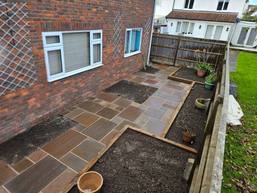 Patio area with stone paving and dark soil plots, bordered by a wooden fence and house.