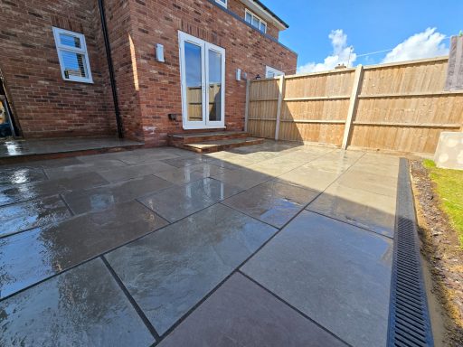 Patio area with wet paving slabs and a wooden fence under a blue sky.