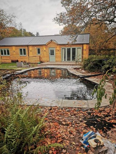 Wooden cabin by a pond surrounded by autumn foliage, grey sky overhead.