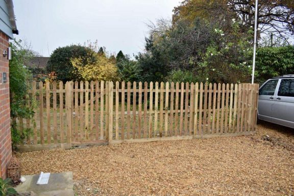 Wooden picket fence surrounding a gravel area with trees and bushes in the background.