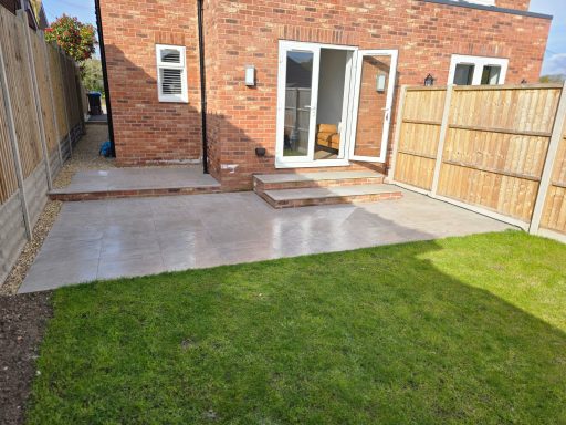 Paved area adjacent to a house, with a small lawn and wooden fence.