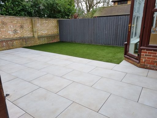 Paved outdoor area with synthetic grass and a brick wall in the background.