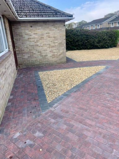 Corner of a brick-paved area with a triangular gravel section and a bushy backdrop.